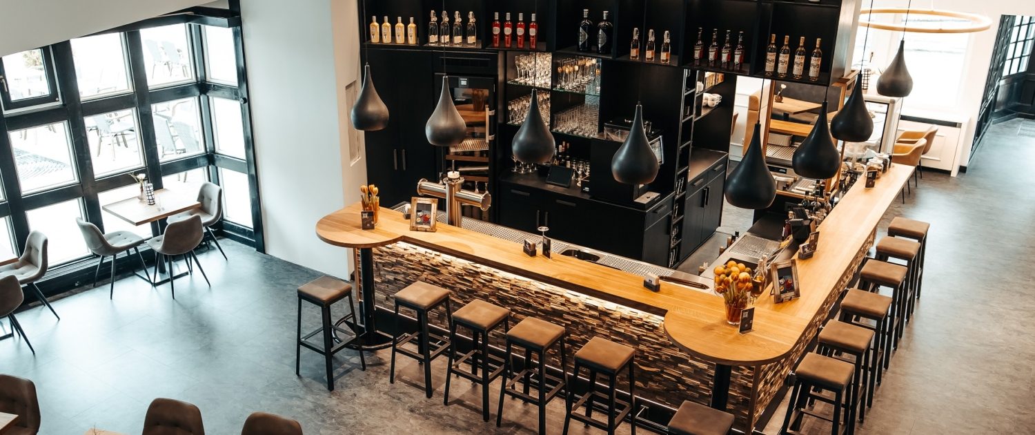 Blick von oben auf die Bar im Mittendrin Café & Lounge des Parkhotel Nümbrecht, mit Holztheke, Barhockern und dekorativer Beleuchtung.