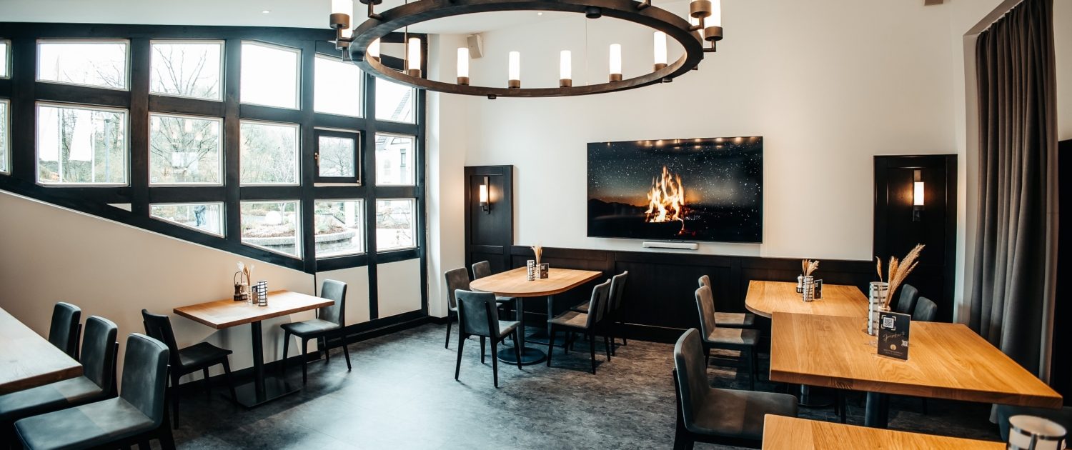 Der Brauhaus-Bereich im Mittendrin Café & Lounge des Parkhotel Nümbrecht mit Holztischen, modernen Stühlen und einem großen Bildschirm mit Kaminfeuer.