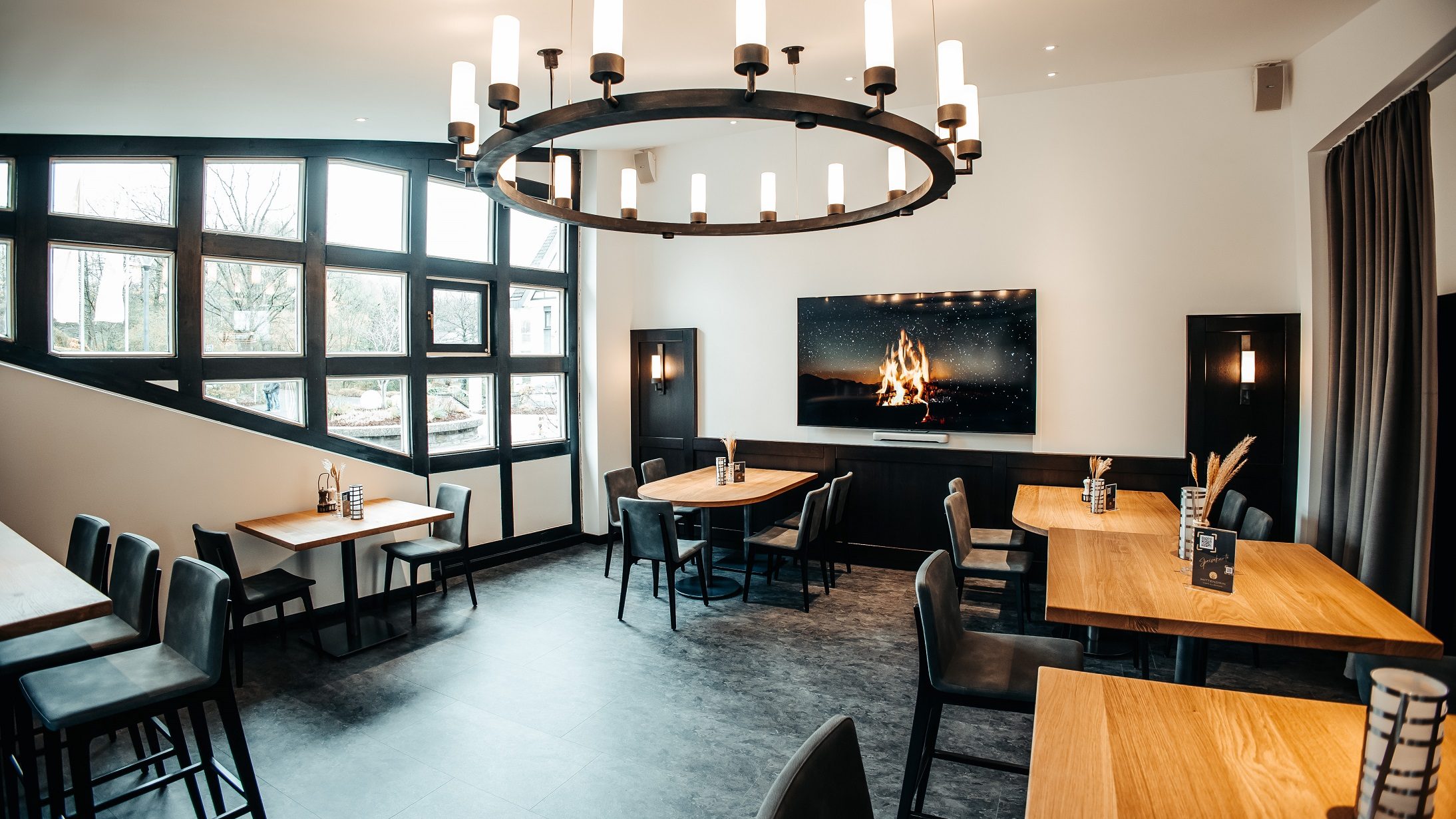 Der Brauhaus-Bereich im Mittendrin Café & Lounge des Parkhotel Nümbrecht mit Holztischen, modernen Stühlen und einem großen Bildschirm mit Kaminfeuer.