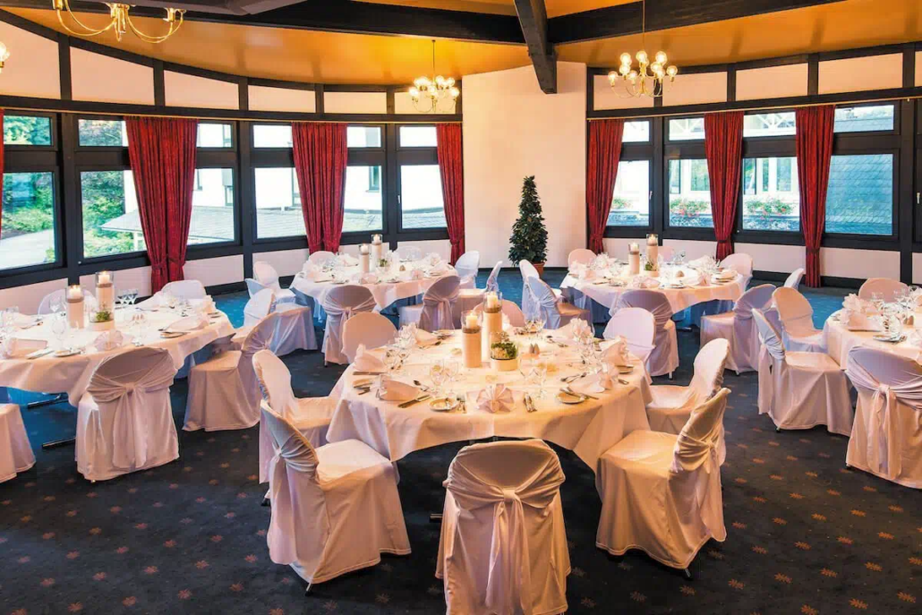 Festlich gedeckter Saal mit runden Tischen, weißen Hussen und Kerzendekoration.