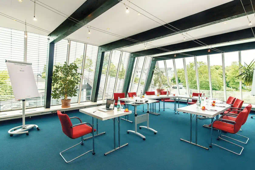 Heller Seminarraum mit großen Fensterfronten, Tischen in U-Form und roten Stühlen.