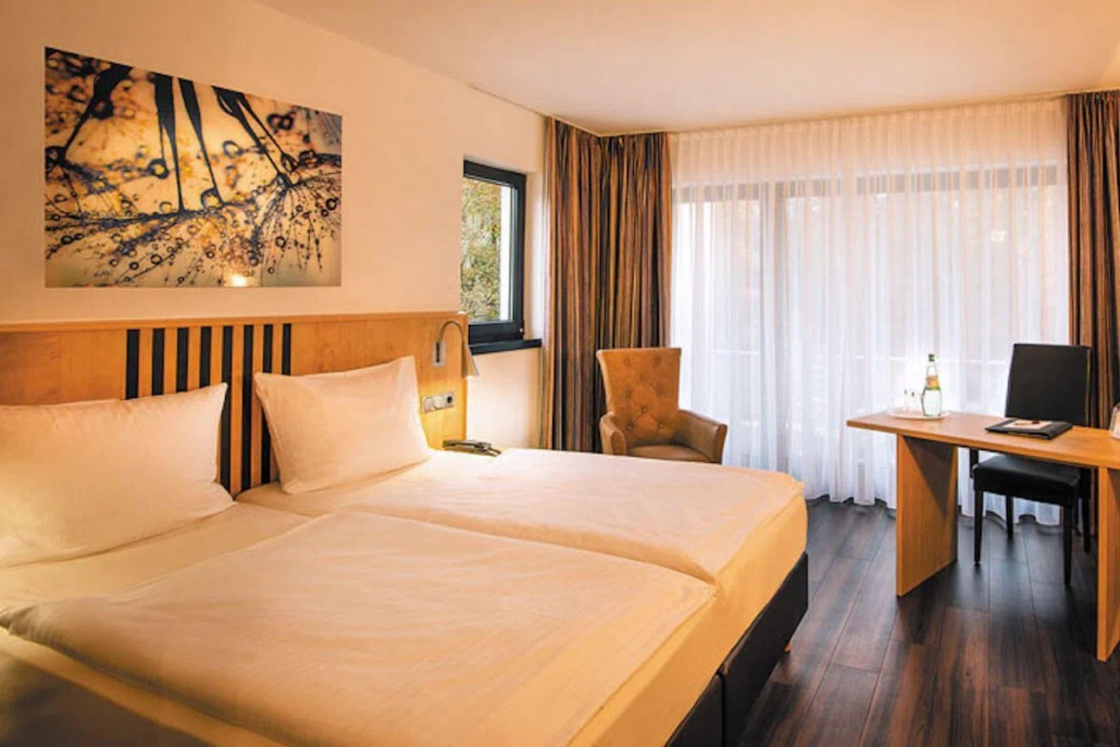 Helles Hotelzimmer mit Doppelbett, Nachttisch und Vorhängen vor dem Fenster/Balkon.