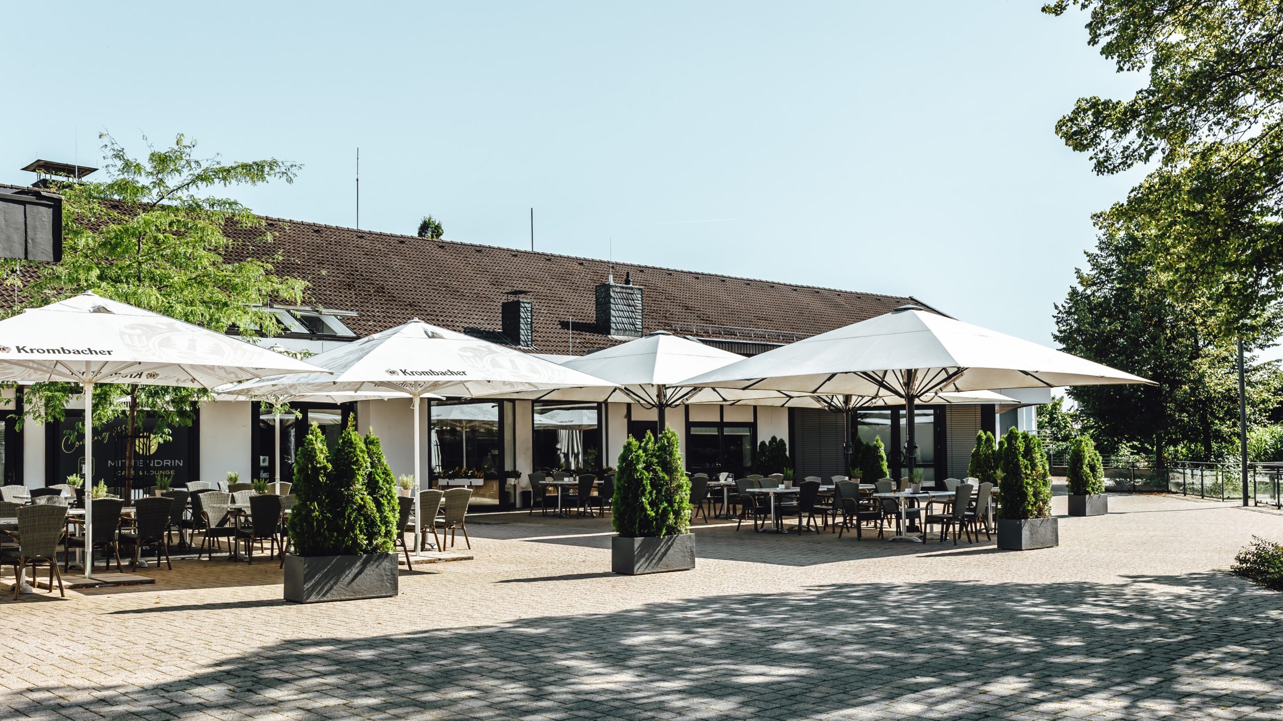Die Weiherterrasse des Mittendrin Café & Lounge im Parkhotel Nümbrecht mit Sonnenschirmen, Tischen und Stühlen an einem sonnigen Tag.