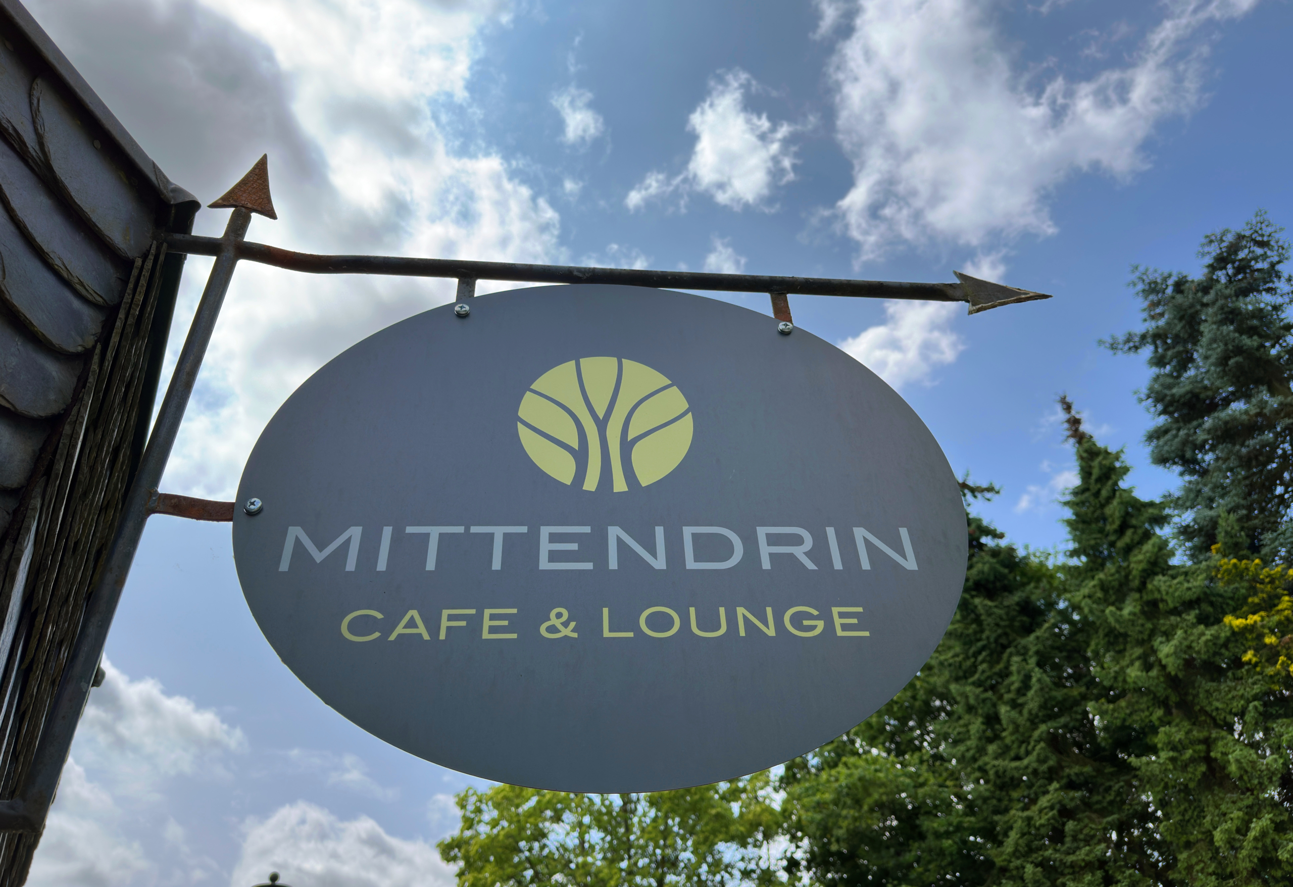 Rundes Schild „Mittendrin Café & Lounge“ vor blauem Himmel.